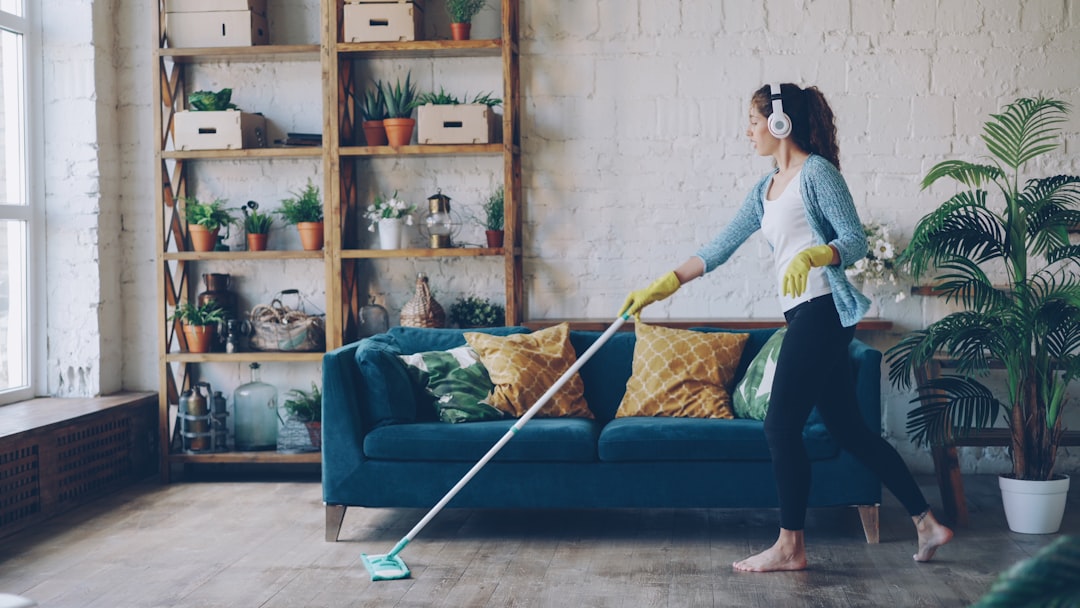 Cheerful young woman is dancing with mop during routine clean-up in nice loft style apartment, she is listening to music with wireless headphones and singing enjoying her occupation.